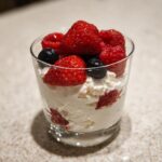 A glass serving of white, fluffy high protein cheesecake fluff topped generously with fresh strawberries, raspberries, and blueberries.