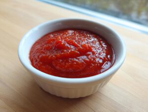 A close-up of vibrant red homemade cocktail sauce recipe served in a small white ramekin on a light wooden surface.