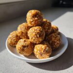 A stack of homemade, golden-brown protein balls coated in visible rolled oats, served on a small white plate.