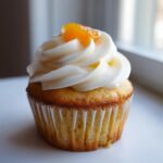 A single, beautifully frosted honey peach cream cheese cupcake topped with a piece of candied peach.