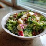 Close-up of a white bowl filled with creamy honeycrisp apple broccoli salad, featuring bright green florets and diced red-skinned apples.