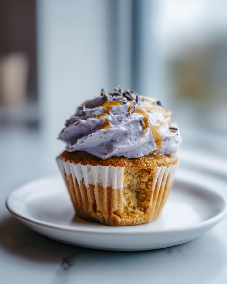 A single lavender honey cupcake with purple frosting, drizzled with honey and topped with lavender buds, sitting on a white plate.