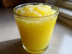 A close-up of a refreshing, bright yellow lemon juice tonic slush served in a clear glass, topped with crushed ice.