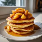 A tall stack of fluffy apple pancakes topped generously with warm, caramelized apple slices and syrup.