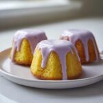 Three vibrant yellow mini lemon cakes with thick, dripping lavender glaze sitting on a white plate.