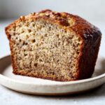 Close-up of a thick slice of moist banana bread for two, showing dark flecks and topped with chopped nuts.