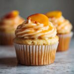 A close-up of a single, moist honey peach cupcake topped with swirled frosting and a slice of fresh peach.