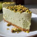 A thick slice of creamy, light green no bake pistachio cheesecake on a white plate, topped with chopped pistachios.