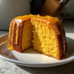 Close-up of a slice cut from a vibrant orange buttermilk cake, topped with a thick orange glaze and zest.