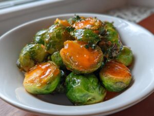 Close-up of vibrant green Orange Maple Glazed Brussels Sprouts coated in a shiny, orange glaze, served in a white bowl.