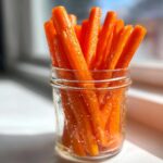 Bright orange, shiny sticks of pickled carrots standing upright in a small glass jar.