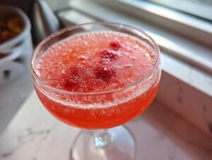Close-up of a bright red raspberry honey lemon daiquiri, garnished with fresh raspberries, served in a coupe glass near a window.