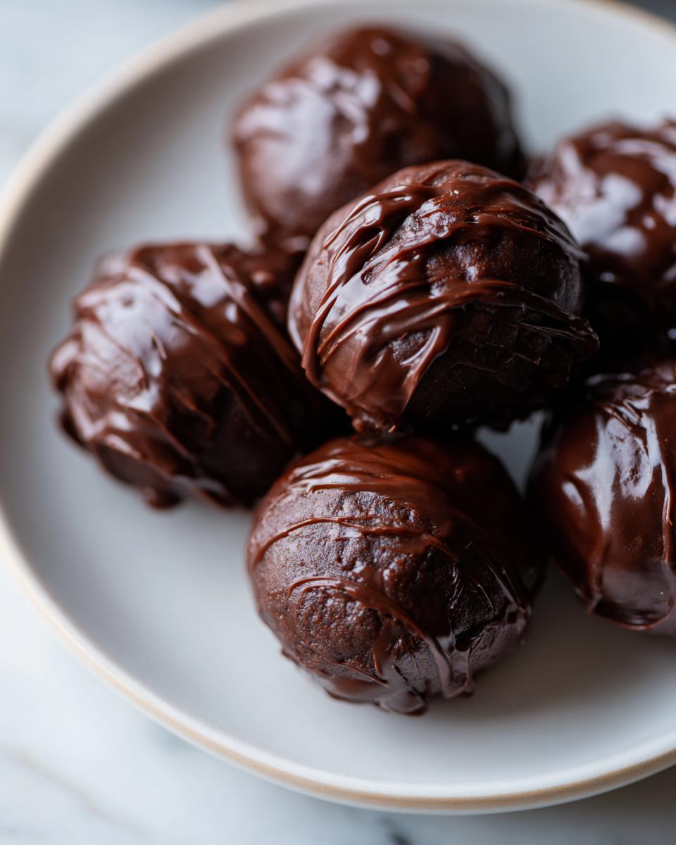 A close-up shot of several decadent, chocolate-covered oreo balls piled on a light-colored plate.