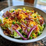 A close-up of the top layer of a seven layer salad featuring bacon, shredded cheese, red onion, and peas.