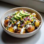 A close-up of loaded southwest potato bowls featuring roasted potatoes, black beans, corn, red onion, avocado slices, and a white drizzle.