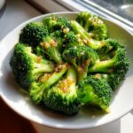 Bright green, perfectly steamed broccoli florets topped with golden brown minced garlic served in a white bowl.