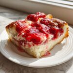 A close-up of a square slice of strawberry cheesecake dump cake on a white plate, topped with bright red strawberry glaze.