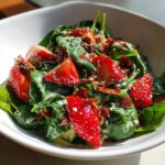A close-up of a vibrant strawberry crackle salad featuring fresh spinach, sliced strawberries, and crispy crackle pieces coated in a creamy dressing.