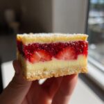 A hand holding a perfect square slice of strawberry custard bars showing three distinct layers: crust, creamy custard, and strawberry topping.