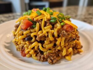 A close-up of a serving of taco pasta salad featuring seasoned ground beef, elbow macaroni, black beans, topped with shredded cheese, diced tomatoes, and green onions.