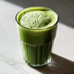 A glass filled with a vibrant green, frothy fat burning smoothie sitting on a light surface.