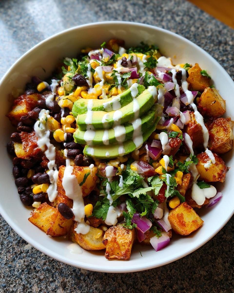 A close-up of a vibrant southwest potato bowls featuring roasted potatoes, black beans, corn, red onion, cilantro, and sliced avocado with a white sauce drizzle.