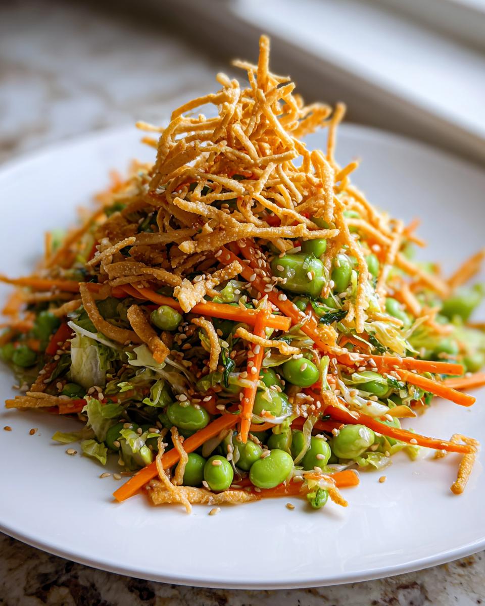 A vibrant Asian edamame salad featuring bright green edamame, shredded carrots, and topped with crispy chow mein noodles and sesame seeds.