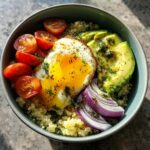 A vibrant avocado egg quinoa bowl featuring a sunny-side-up egg, sliced avocado, cherry tomatoes, and red onion.