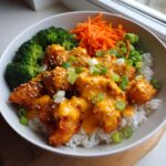 A close-up view of a bang bang chicken bowl featuring crispy chicken coated in orange sauce over white rice, served with broccoli and shredded carrots.