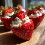 Close-up of bright red, juicy cheesecake stuffed strawberries sitting on a wooden surface in bright light.