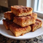 A stack of freshly baked, square churros bars generously coated in cinnamon sugar, resting on a white plate.