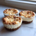 Three delicious mini cheesecakes with a golden brown crumb topping, sitting in white paper liners on a marble surface.