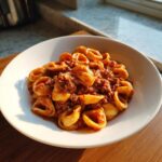 A close-up of a white bowl filled with rich ground beef orecchiette pasta, beautifully lit by natural sunlight.