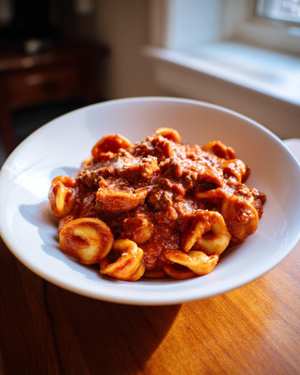 A close-up of a white bowl filled with rich, saucy ground beef orecchiette pasta sitting on a wooden table.