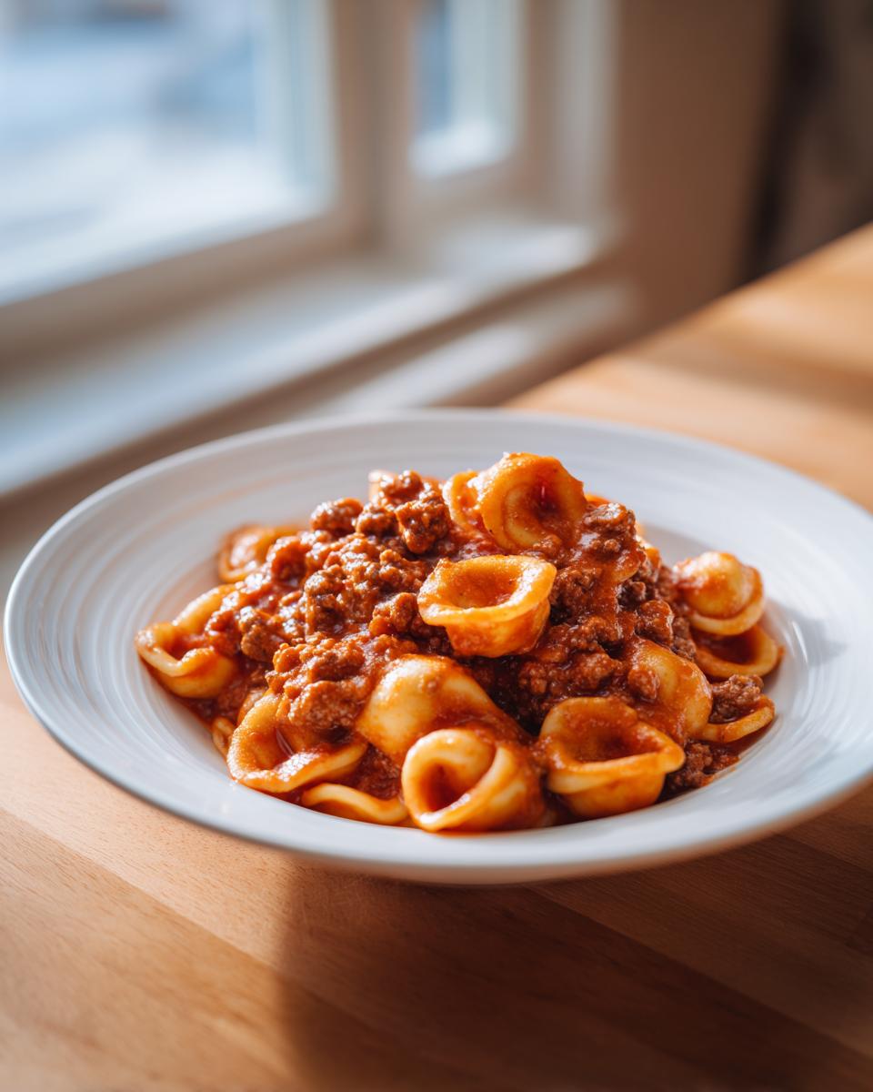 A white bowl filled with delicious ground beef orecchiette pasta coated in rich tomato sauce, sitting on a wooden table.