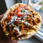 A close-up of a homemade chicken shawarma served open-faced on pita bread, topped with sauce, tomatoes, and red onion.