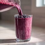 Pouring thick, purple blueberry yogurt smoothie from a blender pitcher into a tall glass topped with fresh blueberries.