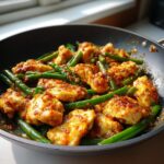 Close-up of juicy chicken pieces coated in a spicy garlic butter sauce mixed with bright green beans in a black skillet.