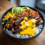 A close-up view of a delicious steak queso rice bowl featuring sliced grilled steak, white rice, bright yellow queso, black beans, corn, salsa, and avocado slices.