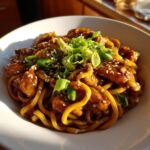 A close-up of glossy, saucy sticky garlic chicken noodles topped with fresh green onions and sesame seeds.