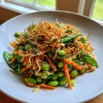 Close-up of a vibrant asian edamame salad featuring bright green edamame, shredded carrots, snow peas, and topped with crispy chow mein noodles and sesame seeds.